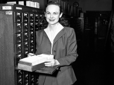 Library staff member Simone Robbins stands beside a card catalog smiling at the camera. A card file rests before her. Photo taken on February 10, 1954.